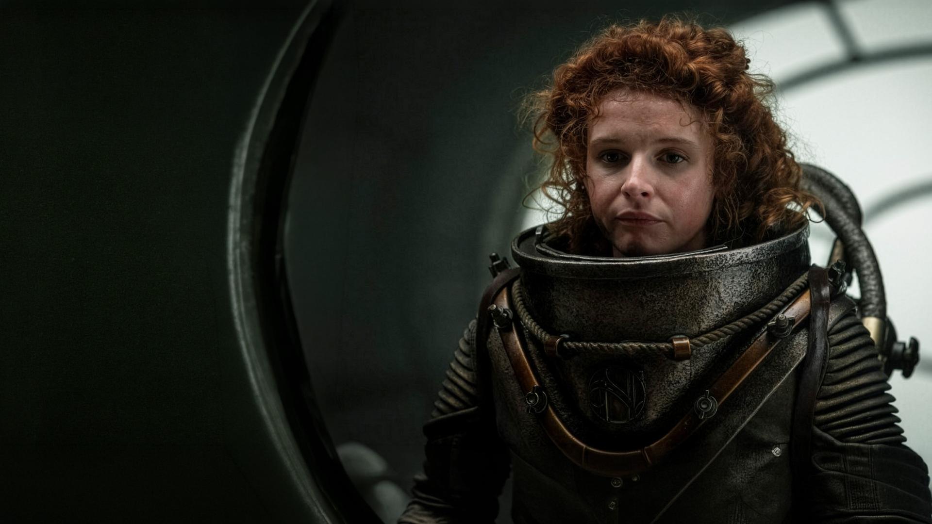 Humility (Georgia Flood) hält sich in einem Raum auf. Sie trägt einen Tiefseetaucheranzug ohne Helm.