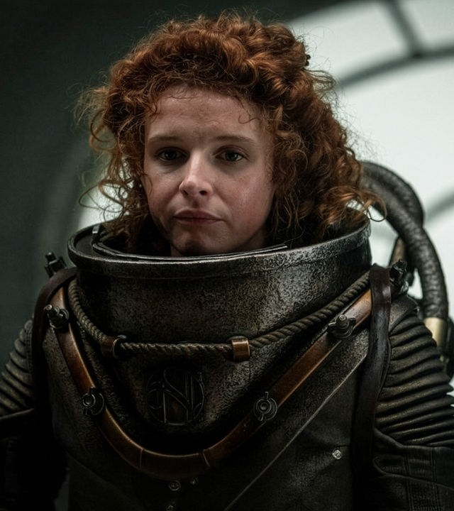 Humility (Georgia Flood) hält sich in einem Raum auf. Sie trägt einen Tiefseetaucheranzug ohne Helm.