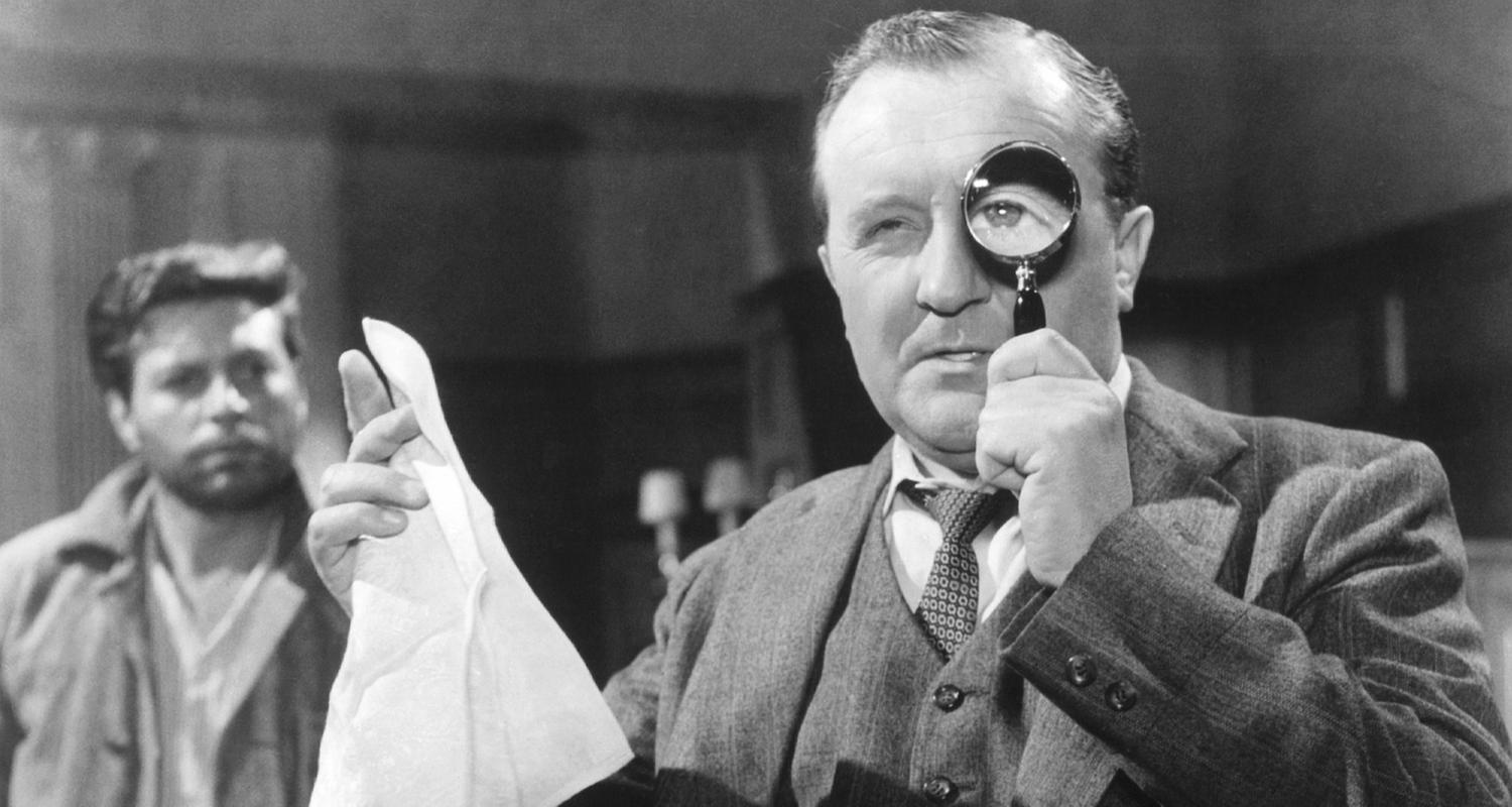 Spielszene in Schwarz-Weiß: Ein Mann im Anzug hält sich eine Lupe vor das linke Auge, in der rechten Hand hält er ein Taschentuch. Im Hintergrund beobachtet ihn ein bärtiger Mann in Hemd und Jacke.