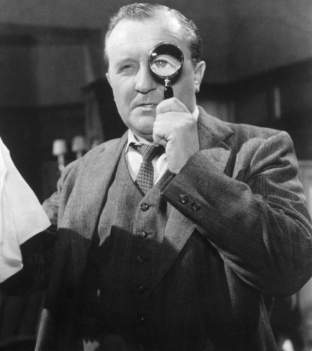 Spielszene in Schwarz-Weiß: Ein Mann im Anzug hält sich eine Lupe vor das linke Auge, in der rechten Hand hält er ein Taschentuch. Im Hintergrund beobachtet ihn ein bärtiger Mann in Hemd und Jacke.