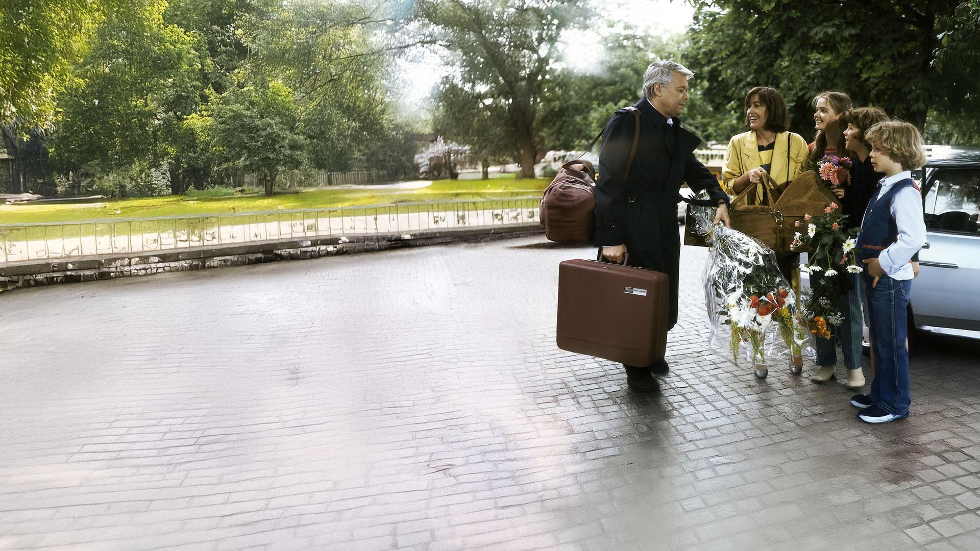 Werner steht auf der Straße seiner Frau Angi und den drei Kindern gegenüber. Er hält einen Blumenstrauß, Angi eine Babytrage in der Hand.