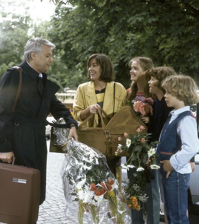 Werner steht auf der Straße seiner Frau Angi und den drei Kindern gegenüber. Er hält einen Blumenstrauß, Angi eine Babytrage in der Hand.
