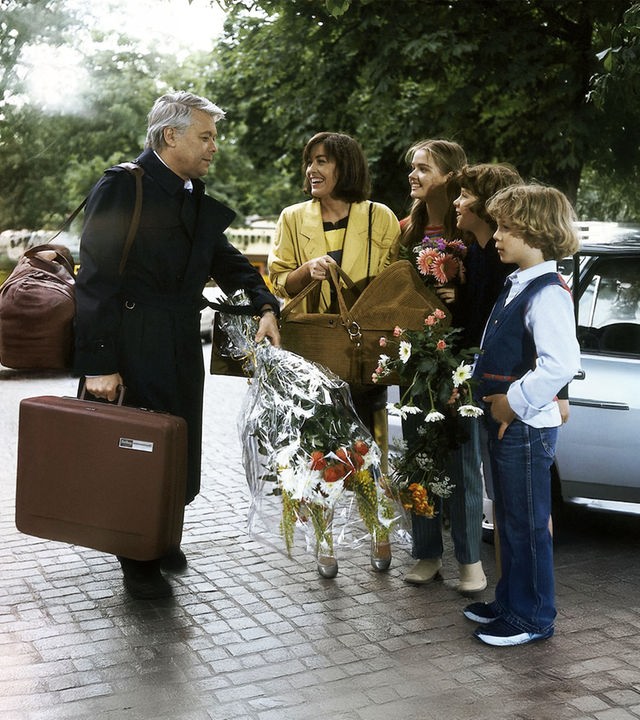 Werner steht auf der Straße seiner Frau Angi und den drei Kindern gegenüber. Er hält einen Blumenstrauß, Angi eine Babytrage in der Hand.