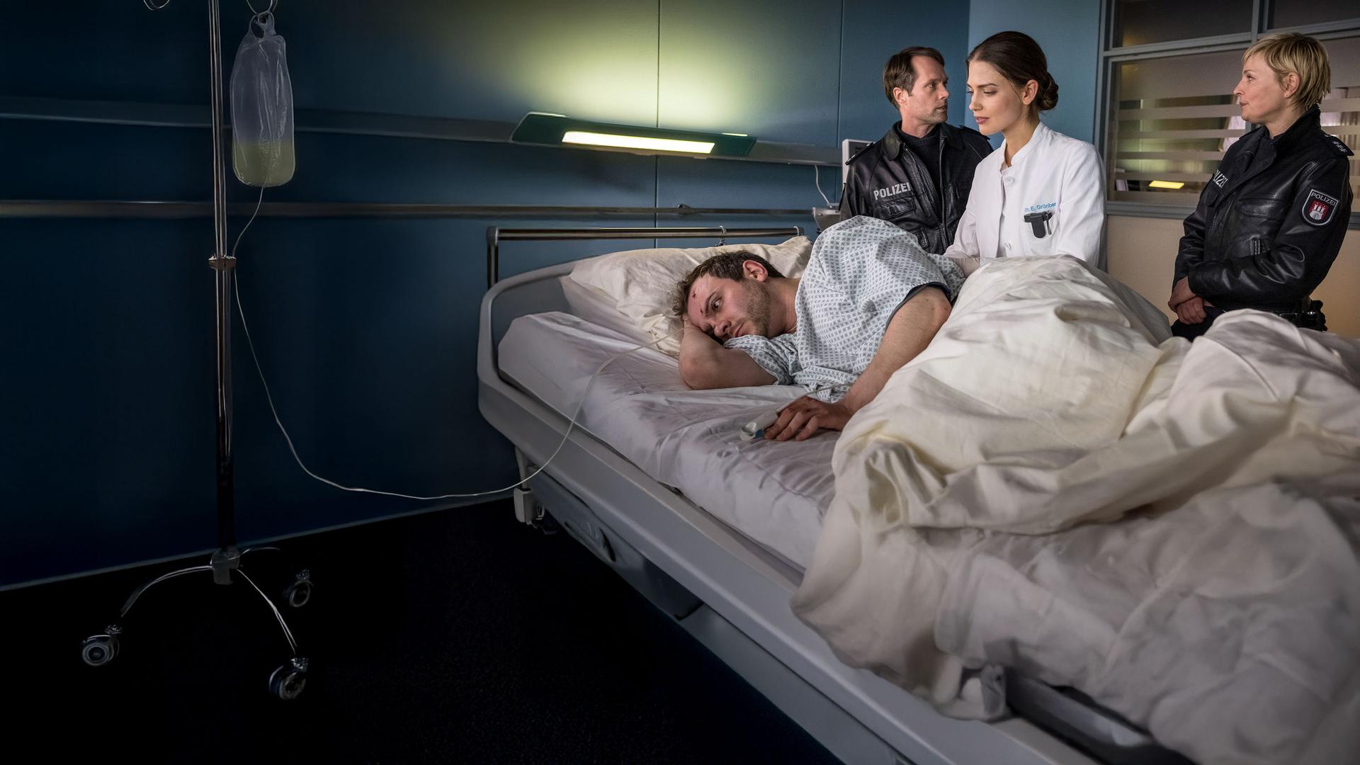 "Notruf Hafenkante - Der große Knall": Die Polizisten Mattes Seeler (Matthias Schloo) und Melanie Hansen (Sanna Englund) stehen auf der rechten Seite des Krankenbetts von Alex Adamy (Eugen Knecht). Zwischen ihnen steht die Ärztin Dr. Eva Grünberg (Laura Berlin). Alex liegt auf der Seite, der Rücken Dr. Grünberg zugewandt.