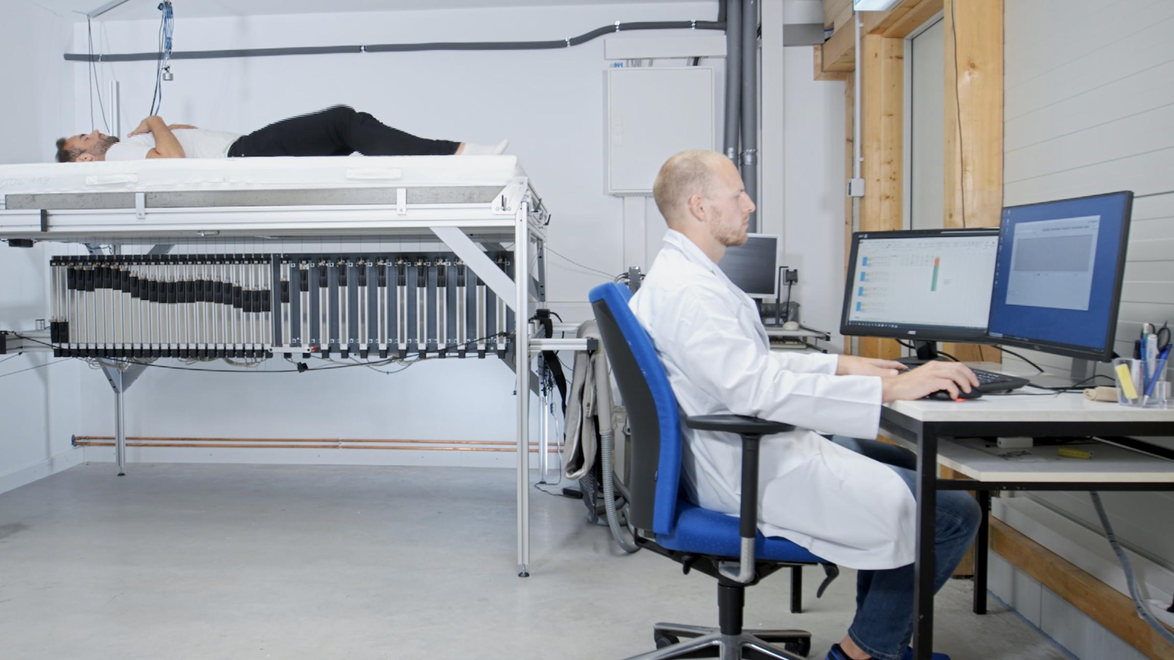 "ZDFzeit: Der große Warentest - Richtig gut schlafen": Ein Mann sitzt im Labor an seinem Schreibtisch. Im Hintergrund liegt ein Mann auf einem hohen Tisch auf einer Matratze.