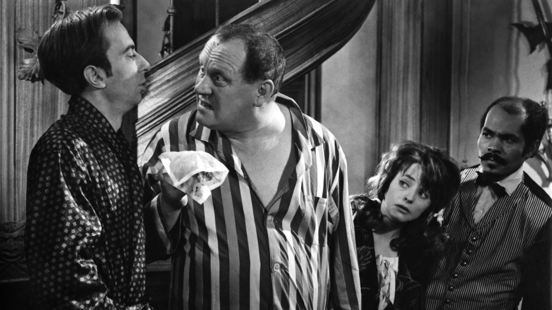 Szenenbild in Schwarz-Weiß: Ein Mann im Pyjama hält einem Mann im Morgenmantel aufgebracht ein blutiges Taschentuch entgegen. Rechts im Hintegrund beobachten ein Mann in Butler-Uniform und eine Frau neugierig das Geschehen.