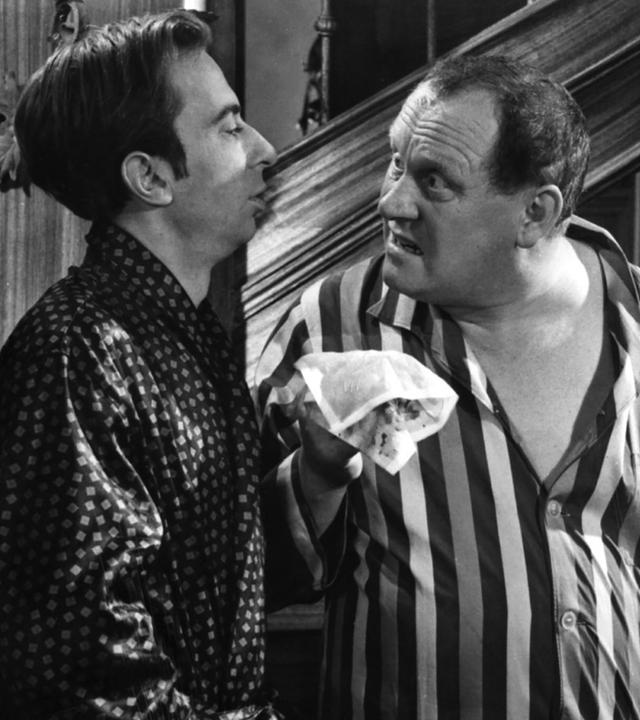 Szenenbild in Schwarz-Weiß: Ein Mann im Pyjama hält einem Mann im Morgenmantel aufgebracht ein blutiges Taschentuch entgegen. Rechts im Hintegrund beobachten ein Mann in Butler-Uniform und eine Frau neugierig das Geschehen.