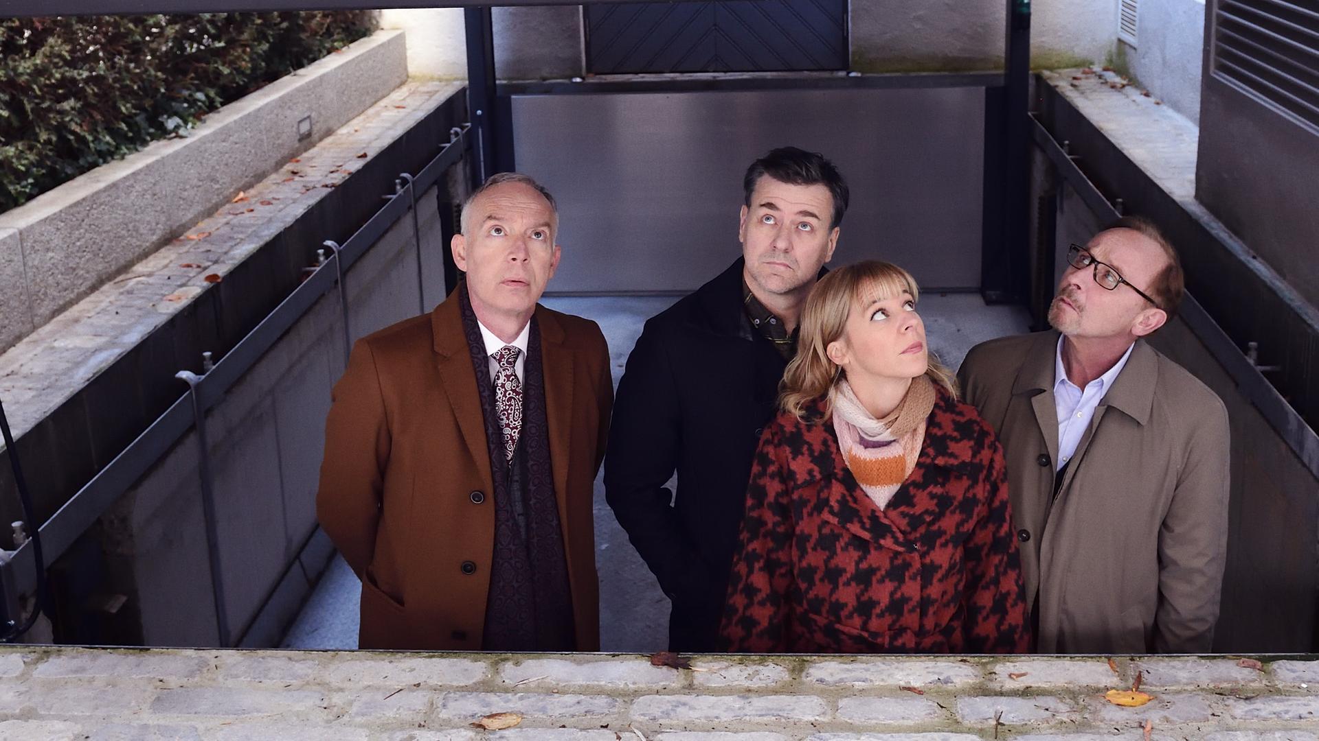 Helmut Zangel (Christoph Süß), Harald Neuhauser (Marcus Mittermeier), Angelika Flierl (Bernadette Heerwagen) und Ludwig Schaller (Alexander Held) blicken aus einem abgesenkten und überdachten Bereich nach oben. Sie sind umgeben von den Wänden einer Villa sowie von einer hohen Hecke.