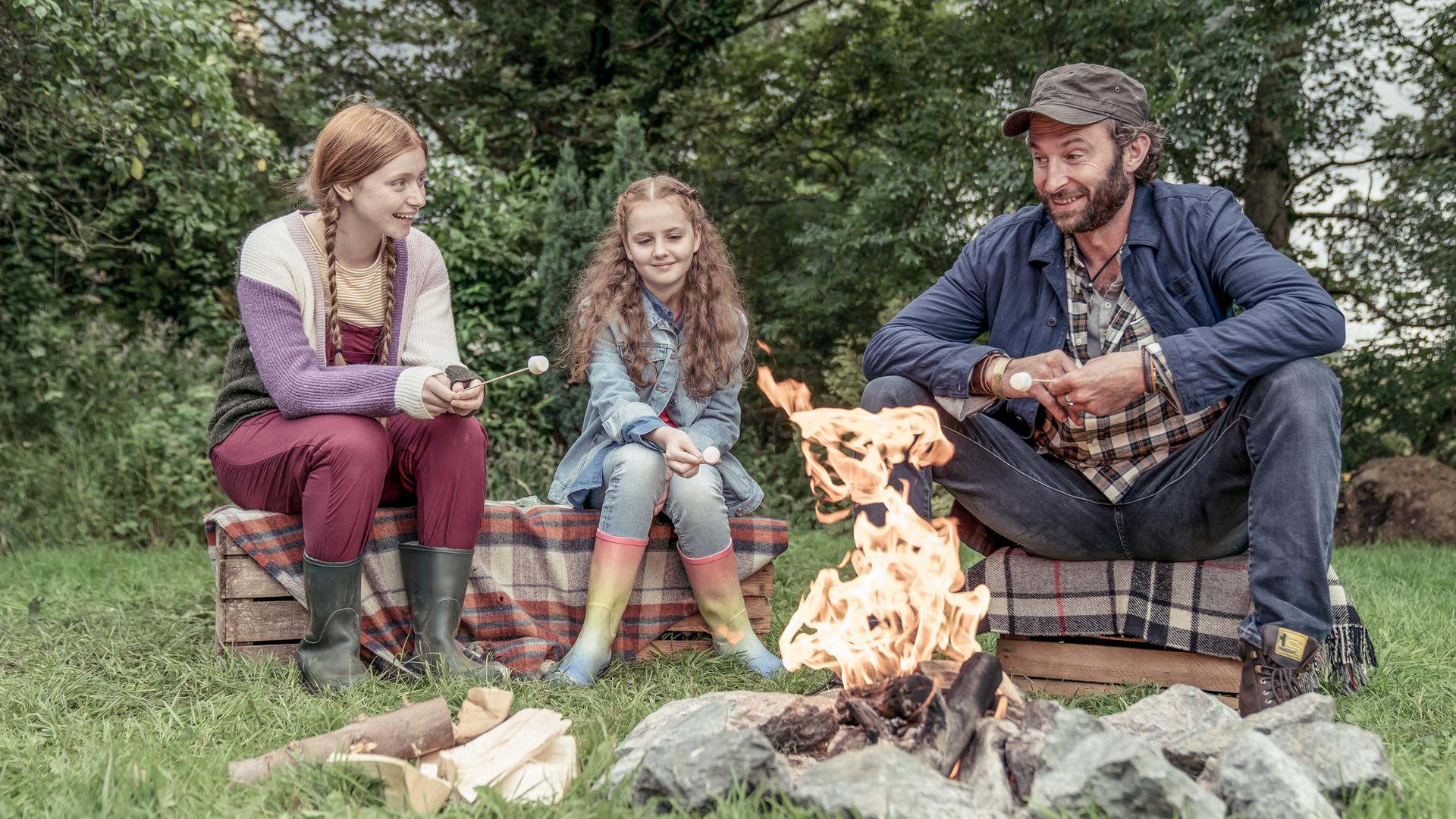 Vor einigen Büschen auf der Wiese an einem Lagerfeuer: Sitzen auf Holzkisten, die mit  karierten Decken überdeckt sind, Mildred, Isabella John und Isabellas Vater. Alle tragen normale Alltagskleidung, lachen und sehen glücklich und entspannt aus.