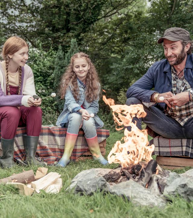Vor einigen Büschen auf der Wiese an einem Lagerfeuer: Sitzen auf Holzkisten, die mit  karierten Decken überdeckt sind, Mildred, Isabella John und Isabellas Vater. Alle tragen normale Alltagskleidung, lachen und sehen glücklich und entspannt aus.