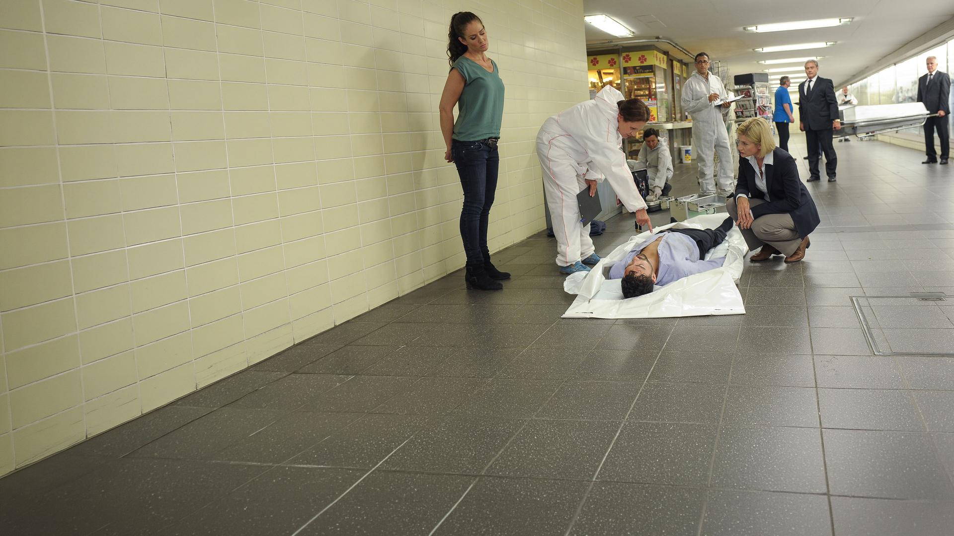 "SOKO Stuttgart - Der Held von Stuttgart": Prof. Lisa Wolter (Eva Maria Bayerwaltes) zeigt Martina Seiffert (Astrid M. Fünderich) etwas auf der Brust des Opfers Daniel Pflüger (Komparse). Selma Kirsch (Yve Burbach) steht daneben und hört zu. Im Hintergrund steht Jan Arnaud (Mike Zaka Sommerfeldt), der die Situation beobachtet sowie Spurensicherer (Komparsen), die die Umgebung absuchen. Die Bestatter (Komparsen) tragen den Sarg zur Leiche.