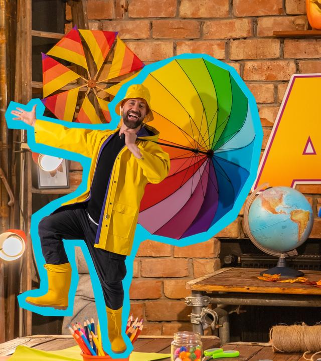 Singa steht vor einem herbstlich dekorierten Hintergrund und zeigt lachend auf einen gelben Papierdrachen, der auf einer Staffelei neben ihr steht. Das SingDing schaut daneben mit weit offenem Mund hinter einem Tisch hervor. Auf dem Tisch steht verkleinert Bürger Lars Dietrich und lacht mit ausgestreckten Armen und Beinen. 