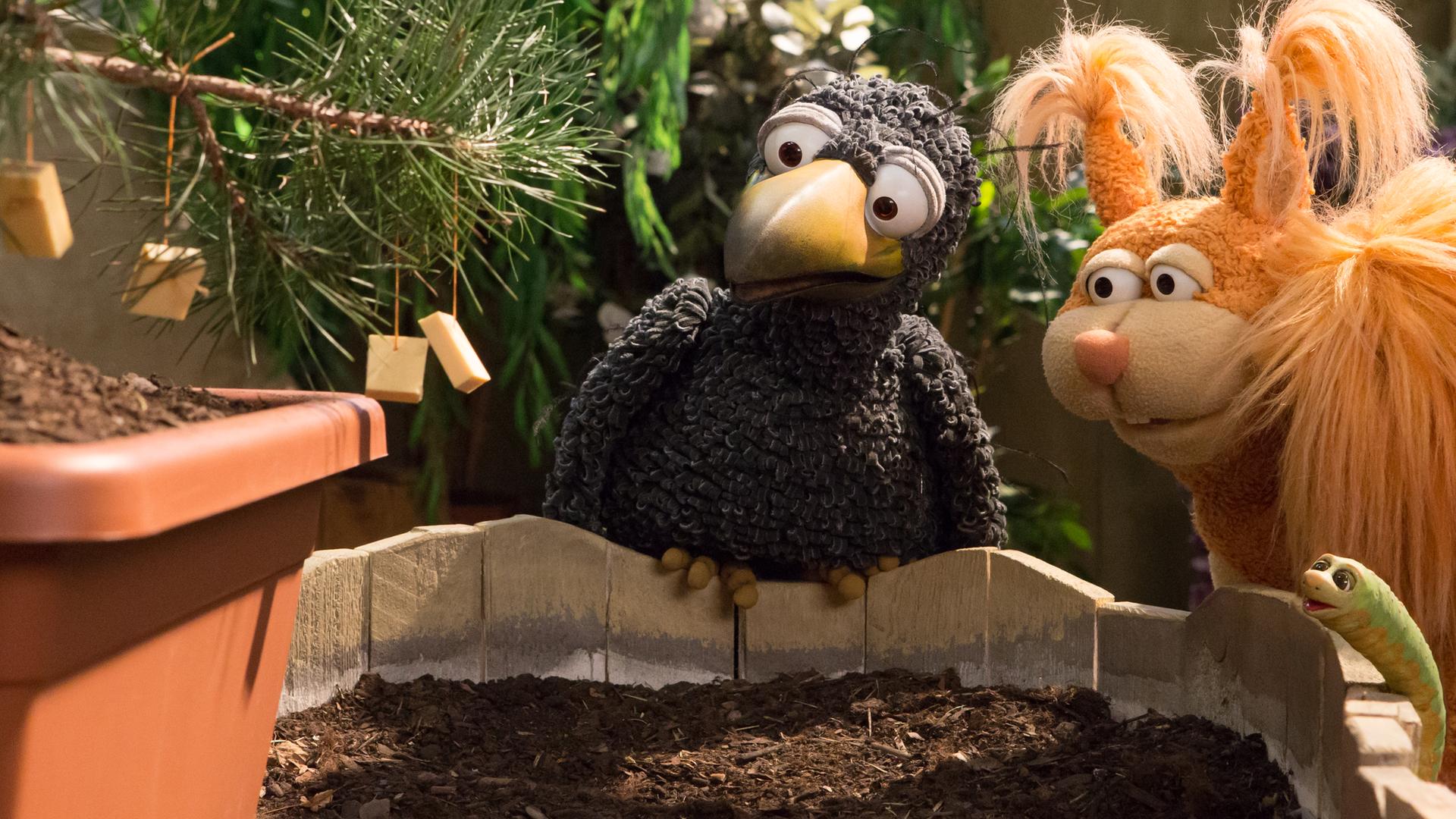 Rudi, Ella und Lotti hocken vor einem Pflanzenkübel. Aus einem Topf wächst ein kleiner Nadelbaum, an dessen Ästen Käsestücke hängen.