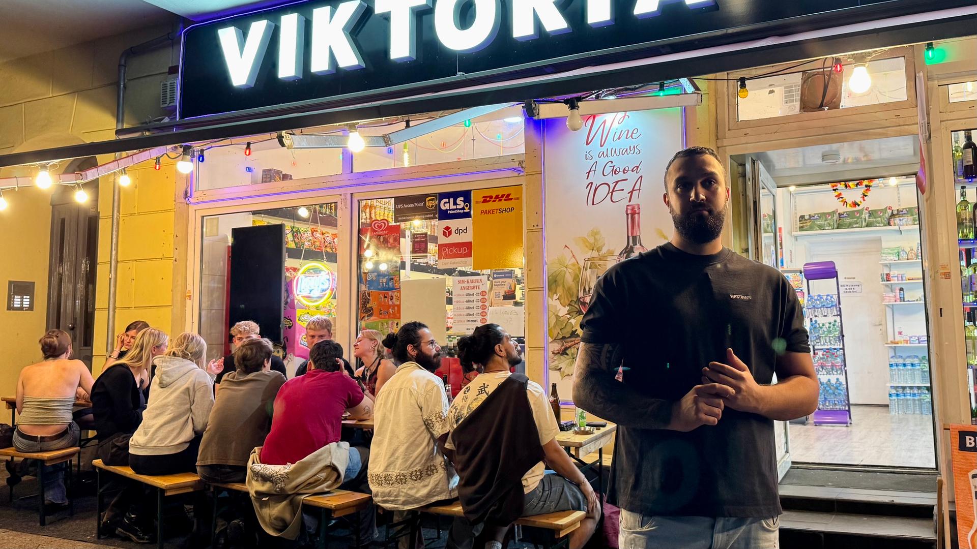 Serdar steht spät in der Nacht vor seinem Kiosk. Im Hintergrund sitzen zahlreiche Gäste auf Bänken, vertieft in Gespräche.