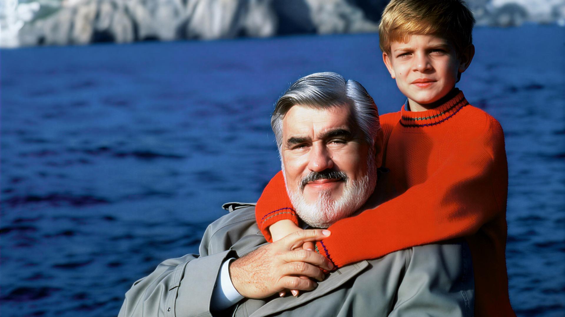 "Der kleine Lord": Carl Schneibel (Mario Adorf) wird liebevoll von Christian (Francesco De Pasquale) umarmt, der hinter ihm steht. Im Hintergrund sind das Meer und Felsen zu sehen.