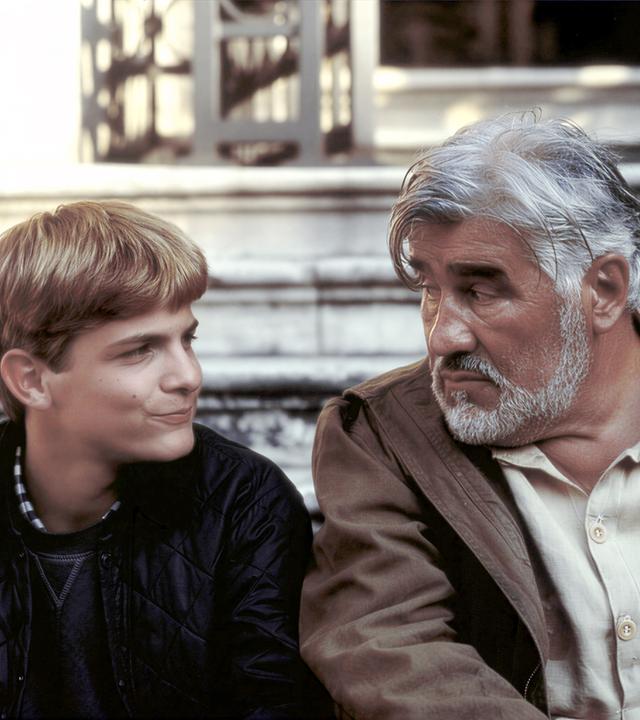"Der kleine Lord - Retter in der Not": Christian (Francesco De Pasquale) und Carl Schneibel (Mario Adorf) sitzen nebeneinander vor einer Treppe und schauen sich gegenseitig an.