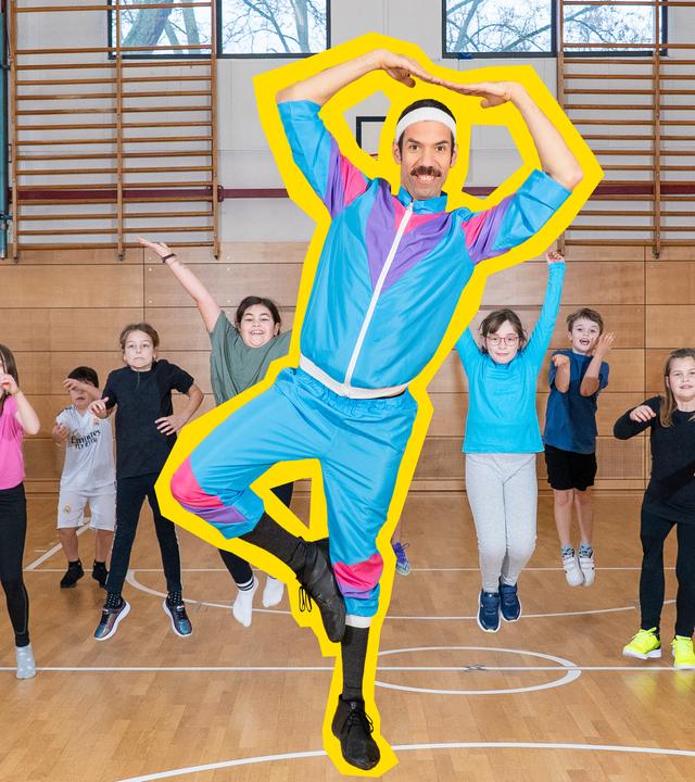  Johannes Stankowski steht auf einem Bein in einer Sporthalle. Die Arme hält er über den Kopf, seine Fingerspitzen berühren sich. Er trägt einen bunten Fitness-Anzug und ein Schweißband und lächelt in die Kamera. Hinter ihm springen 12 Kinder in Sportkleidung auf und ab.