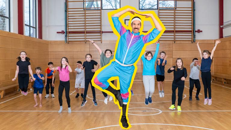  Johannes Stankowski steht auf einem Bein in einer Sporthalle. Die Arme hält er über den Kopf, seine Fingerspitzen berühren sich. Er trägt einen bunten Fitness-Anzug und ein Schweißband und lächelt in die Kamera. Hinter ihm springen 12 Kinder in Sportkleidung auf und ab.