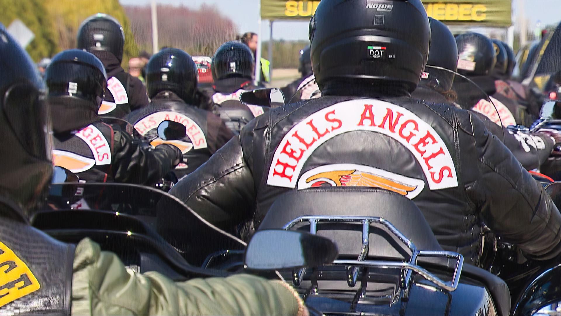 Viele Motorradfahrer mit Lederjacken, auf denen "Hells Angels" steht, fahren auf einer Straße weg von der Kamera.