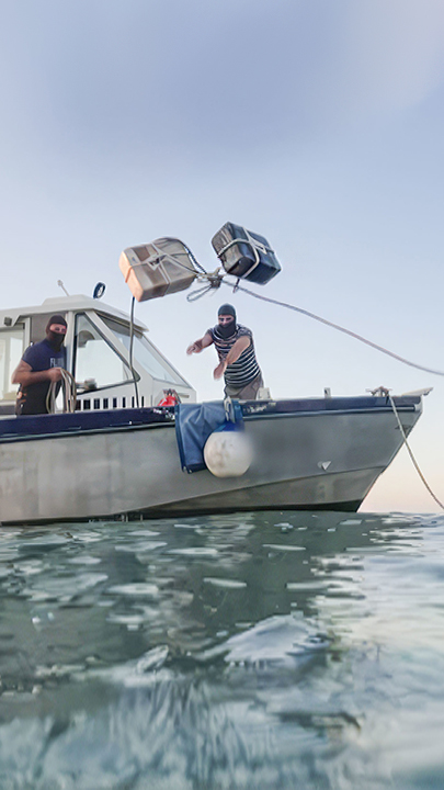 Zwei maskierte Männer werfen zwei Pakete an einem Seil von einem kleinen Boot.