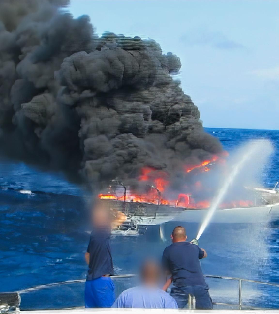 Ein Segelschiff auf hoher See steht in Flammen. Männer löschen es mit einem Schlauch von einem anderen Schiff aus.