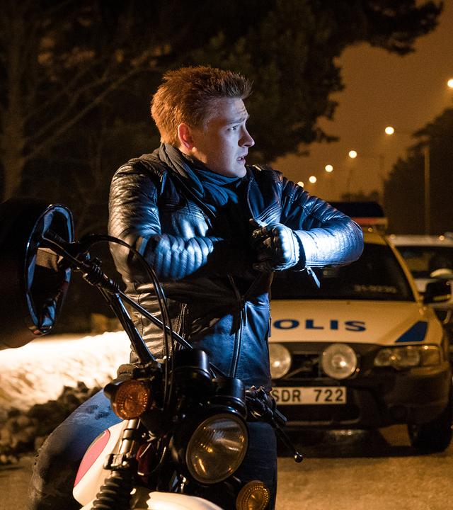 Mats Jörgenson sitzt besorgt schauend auf seinem Motorrad und greift in die linke Brusttasche seiner Lederjacke. Ihm rechts gegenüber steht ein Polizist, der ihn scheinbar angehalten hat. Beide stehen an einem leicht schneebedeckten Straßenrand. Im Hintergrund steht ein Polizeiwagen. Dahinter ist ein weiteres Auto erkennbar.