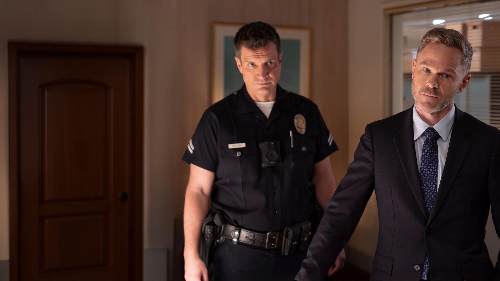 "The Rookie - Der Kuss": Officer John Nolan (Nathan Fillion) und Wesley Evers (Shawn Ashmore) stehen nebeneinander in einem Büroraum. Nolan trägt eine dunkle, kurzärmlige Dienstuniform und blickt ernst nach vorn. Wesley trägt einen schwarzen Anzug und schaut nachdenklich zur Seite.