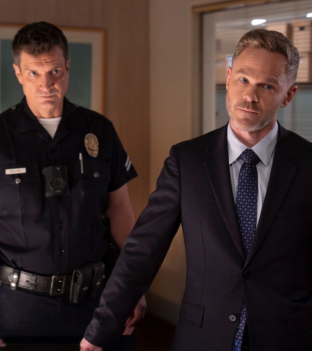 "The Rookie - Der Kuss": Officer John Nolan (Nathan Fillion) und Wesley Evers (Shawn Ashmore) stehen nebeneinander in einem Büroraum. Nolan trägt eine dunkle, kurzärmlige Dienstuniform und blickt ernst nach vorn. Wesley trägt einen schwarzen Anzug und schaut nachdenklich zur Seite.