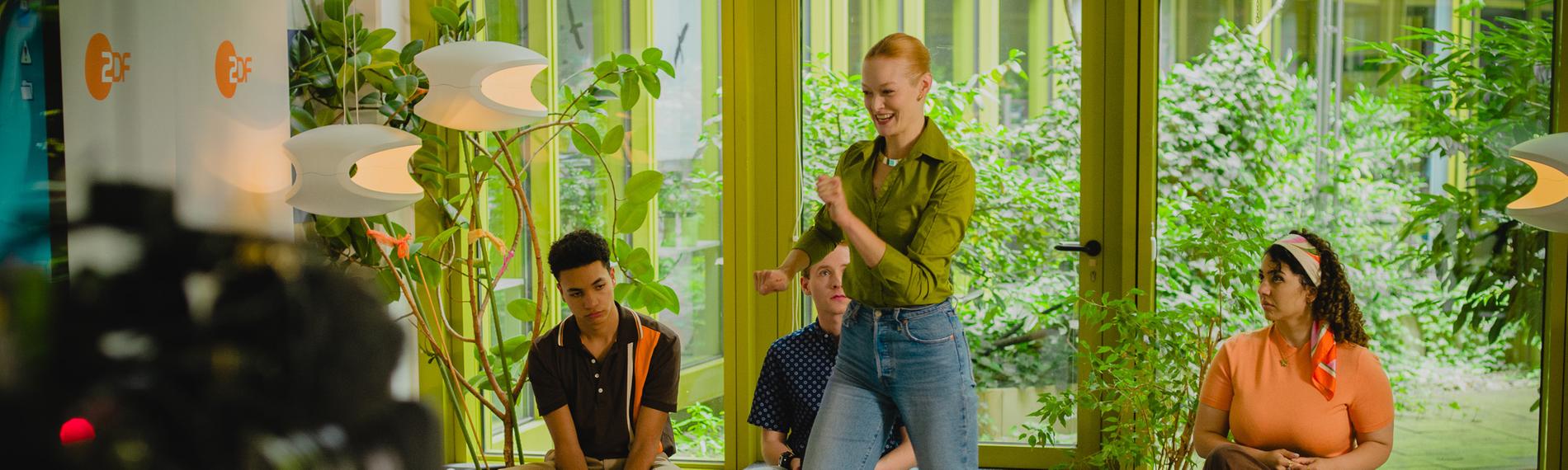 Szenenfoto des Kurzfilms der ZDF-Fiction-Volos: Eine der Volobewerberinnen rennt von ihrem Platz weg, die anderen schauen auf ihren Hockern verblüfft zu. Im Hintergrund Grünplanzen in einem Innenhof.