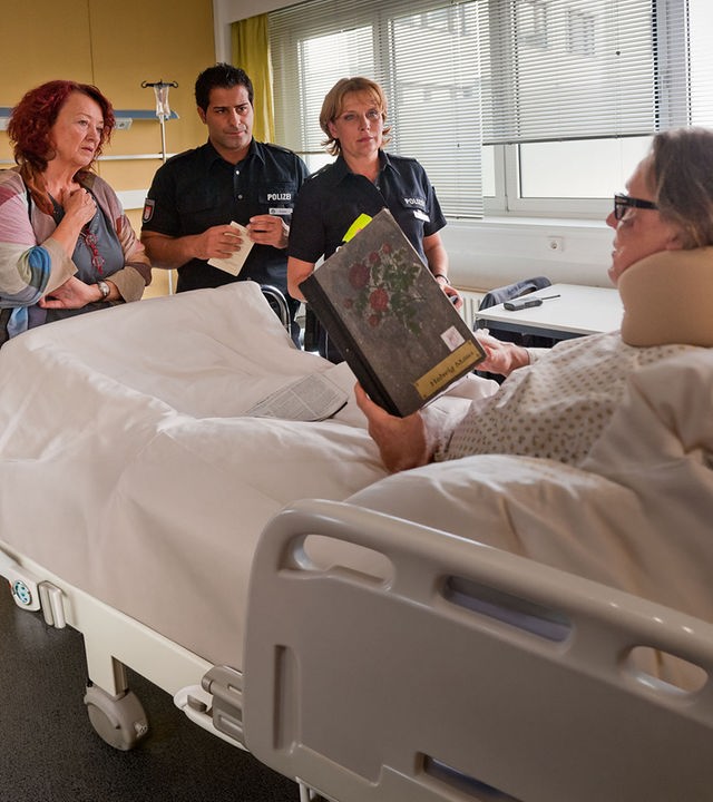 "Notruf Hafenkante - Der letzte Vorhang": Theaterregisseur Harting (Hannes Hellmann) liegt im Krankenhausbett und hält ein Buch in der Hand. Am Fußende des Bettes stehen die Souffleuse Maus (Barbara Focke) sowie die Polizisten Claudia (Janette Rauch) und Tarik (Serhat Cokgezen).