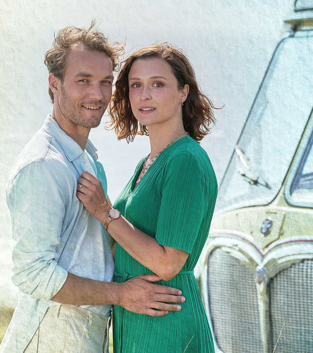 Nicolas (Max Woelky) und Olivia (Isabella Krieger) stehen im Dünengras am Meer und halten sich gegenseitig im Arm. Rechts neben ihnen ist ein grüner Vintage-Bus zu sehen.