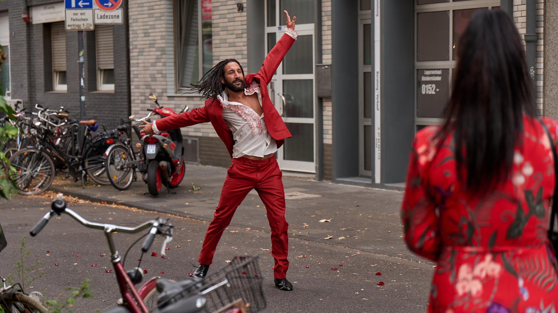 "Heldt - Der Mann aus Wien": Jason Schmoller (Massimo Sinato) steht in Tanzpose mit ausgestreckten Armen auf der Straße. Er trägt einen roten Anzug mit weißem Hemd. Ihm gegenüber steht mit dem Rücken zur Kamera eine Passantin (Komparsin) im roten Kleid, die ihm zuschaut.