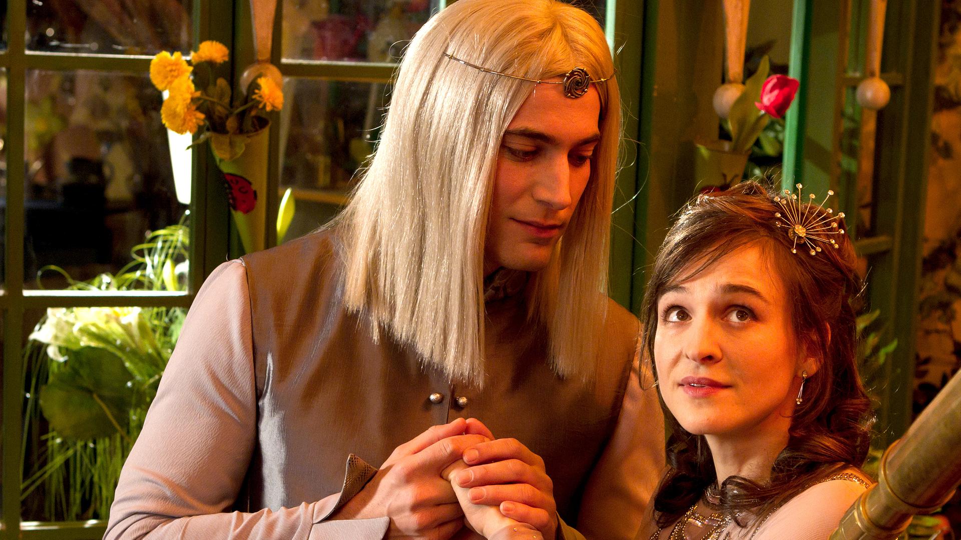 Der Mann im Mond hält die Hände von Siebenstein, die wie eine Prinzessin gekleidet ist und in den Nachthimmel schaut.