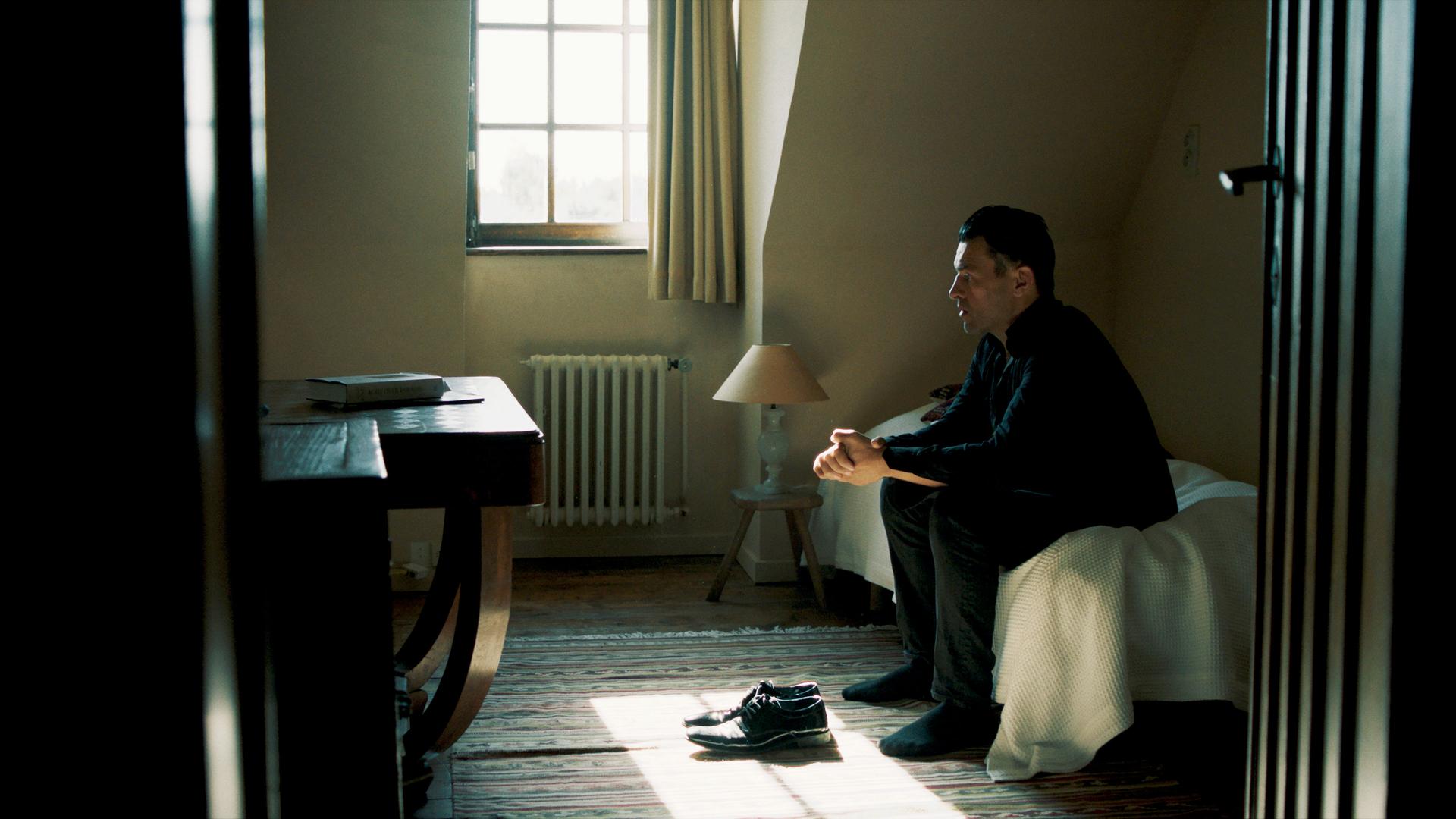 Ein Mann sitzt in einem Zimmer auf dem Bett. Seine Schuhe stehen vor ihm, und er hat die Hände gefaltet.