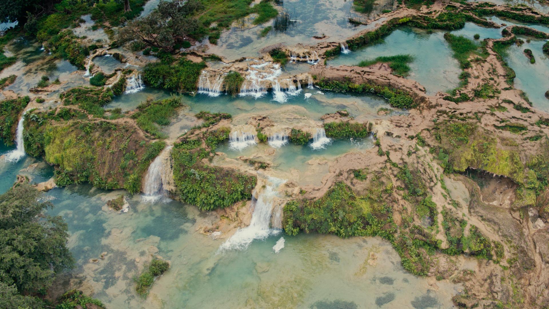 Blick von oben auf kleinere Wasserfälle, die umgeben sind von grünen Flächen.