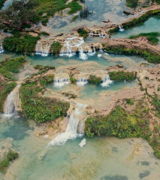 Blick von oben auf kleinere Wasserfälle, die umgeben sind von grünen Flächen.