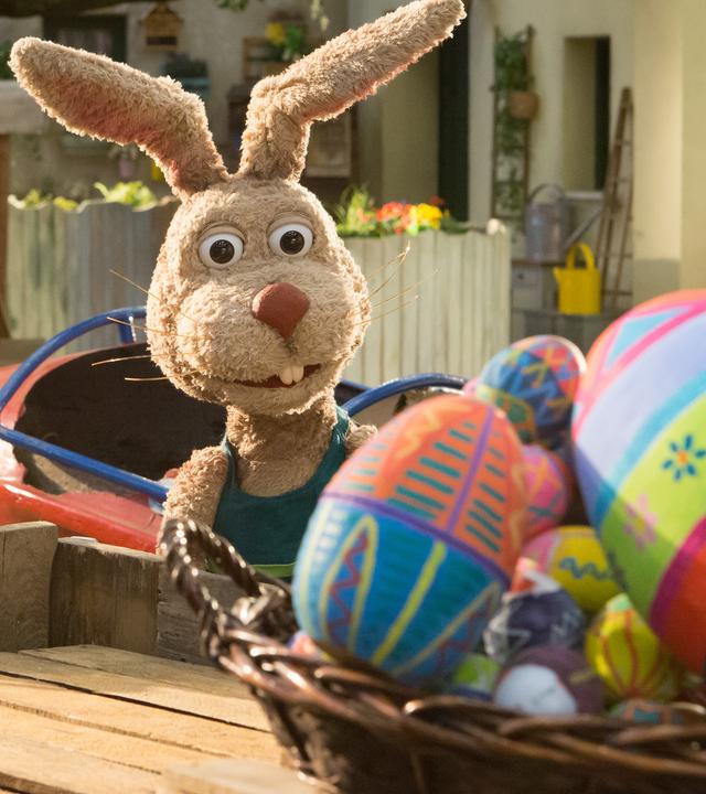 Grete, die ein Körbchen trägt, und Otto schauen erstaunt auf einen Korb voller bunter Ostereier.