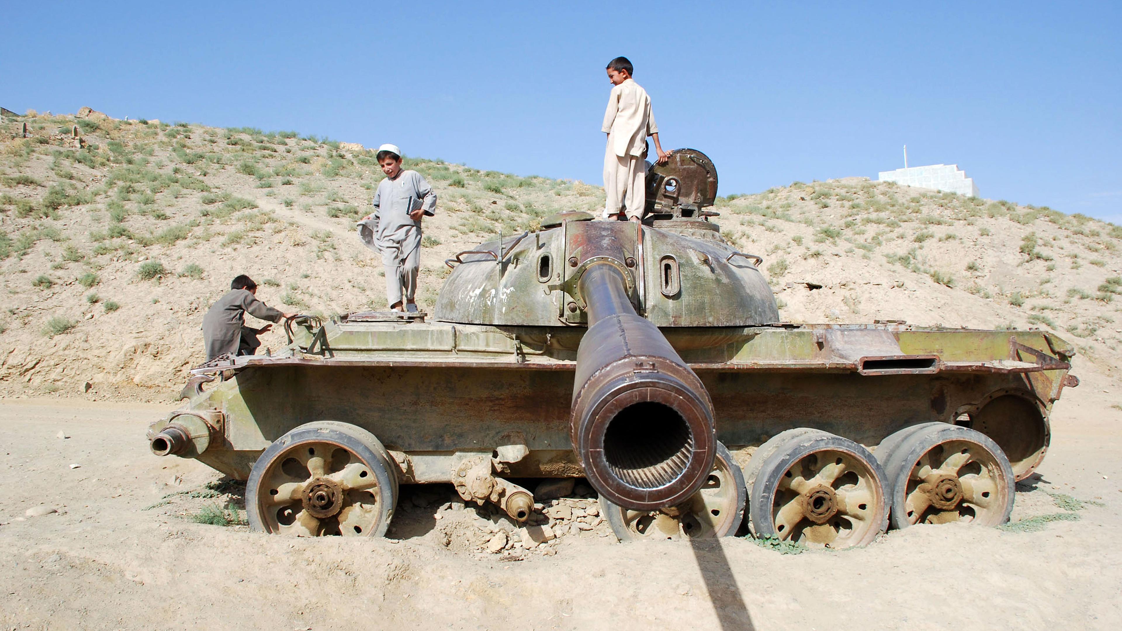 In einer sandigen Landschaft stehen afghanische Kinder auf einem Militärpanzer, das Panzerrohr "zielt" Richtung Kamera.
