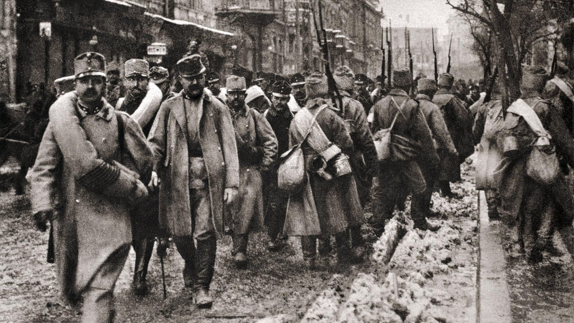 "Der Preis des Krieges - Erster Weltkrieg": Schwarz-Weiß-Fotoaufnahme mit bewaffneten Soldaten marschierend auf einer Straße. Einige blicken nach vorne, andere kehren dem Betrachter den Rücken zu.