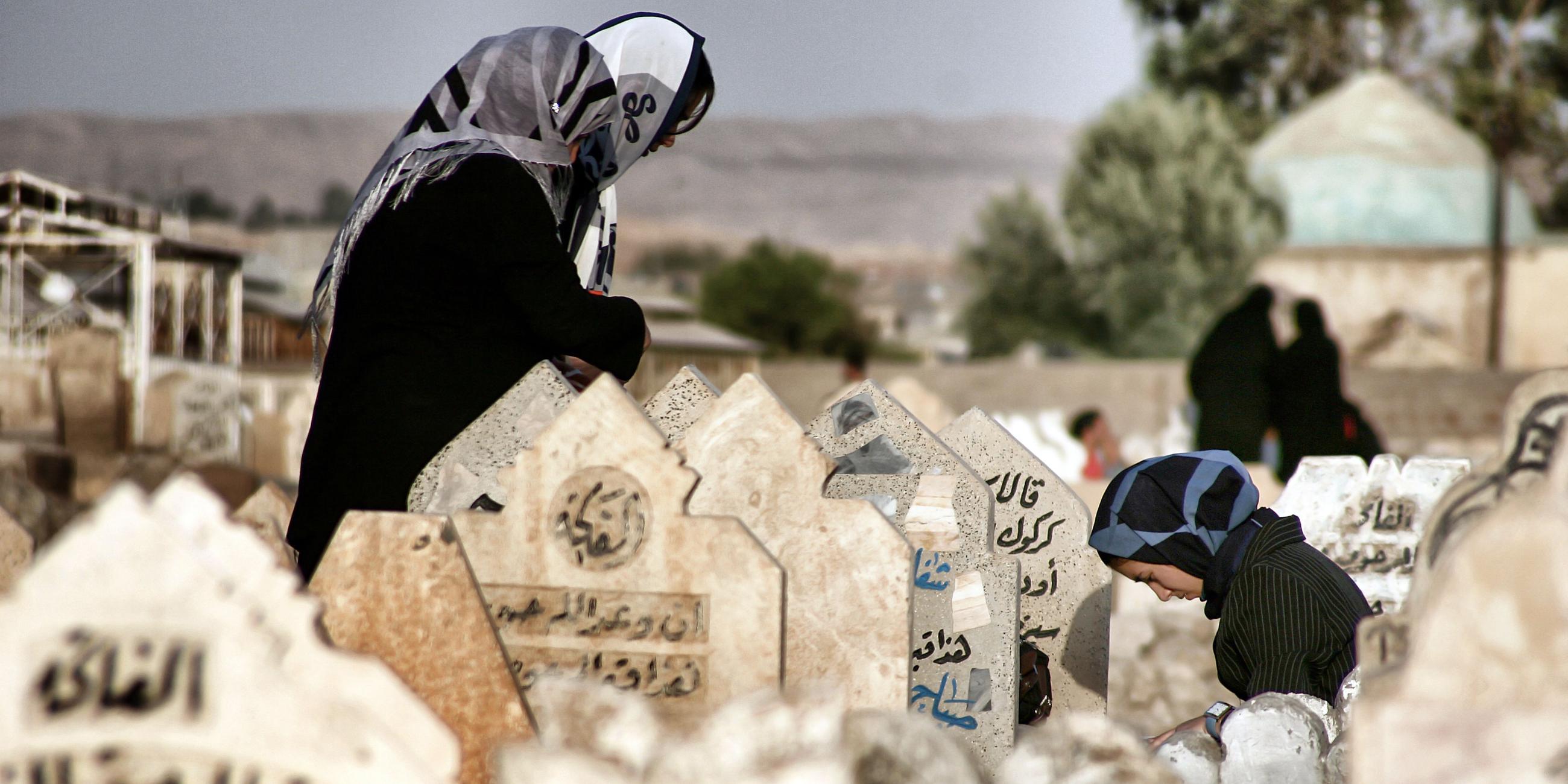 "Der Preis des Krieges: Irak": Drei Frauen mit Kopftüchern stehen mit geneigten Köpfen um ein Grab auf einem Friedhof. Eine Frau kniet. Es sind arabische Inschriften auf den Grabsteinen zu lesen.