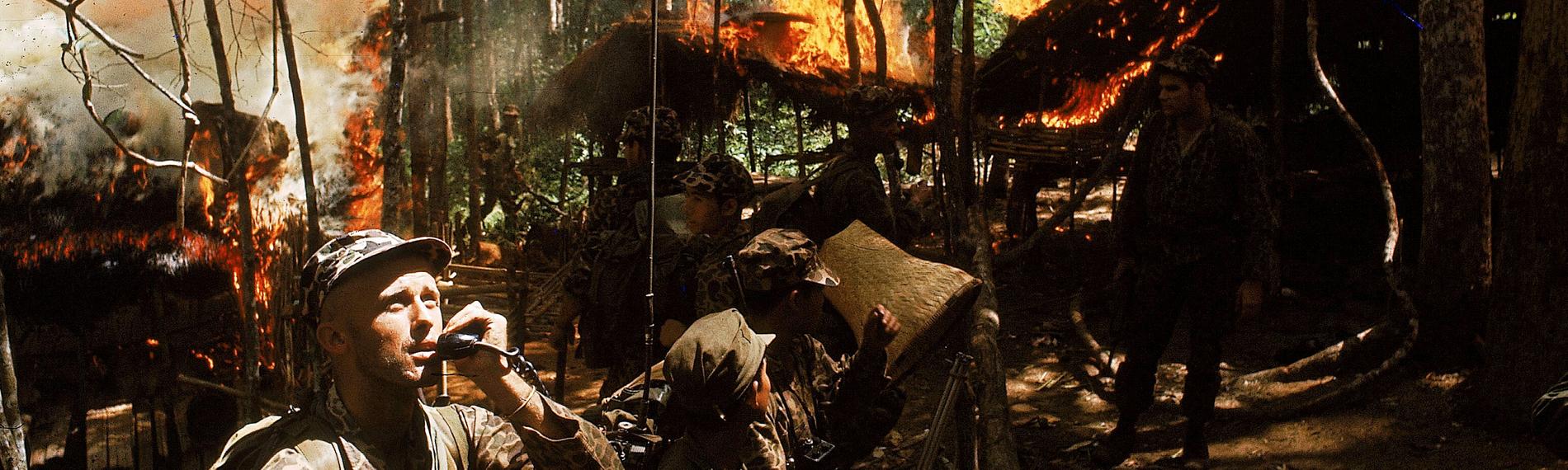 "Der Preis des Krieges - Vietnam": Ein US-Soldat telefoniert via Funk in einem Wald, umgeben von brennenden Hütten. Um ihn herum stehen weitere Soldaten.