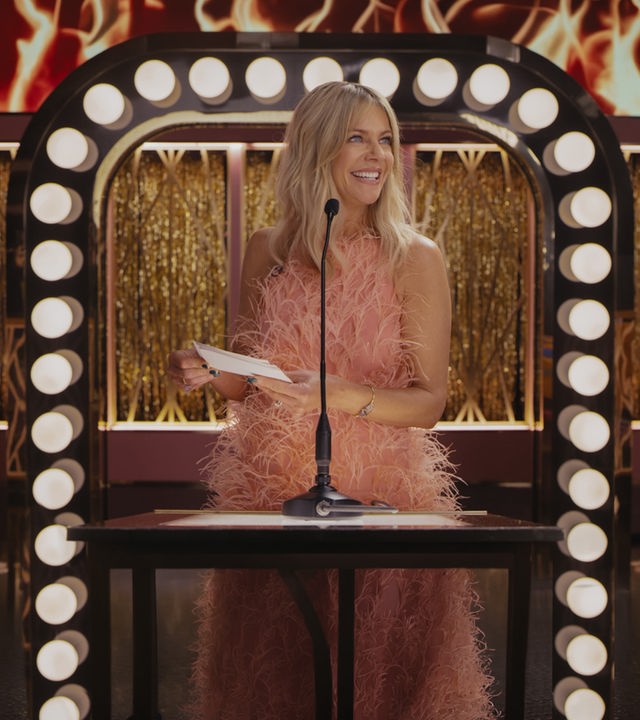 "Hacks - Der Roast der Deborah Vance": DJ (Kaitlin Olson) steht in einem lachsfarbenen, ärmellosen federartig anmutenden Kleid lächelnd vor einem Mikrofon und hält in beiden Händen einen Zettel. Im Hintergrund sind die Lichter der Bühnendekoration zu sehen.
