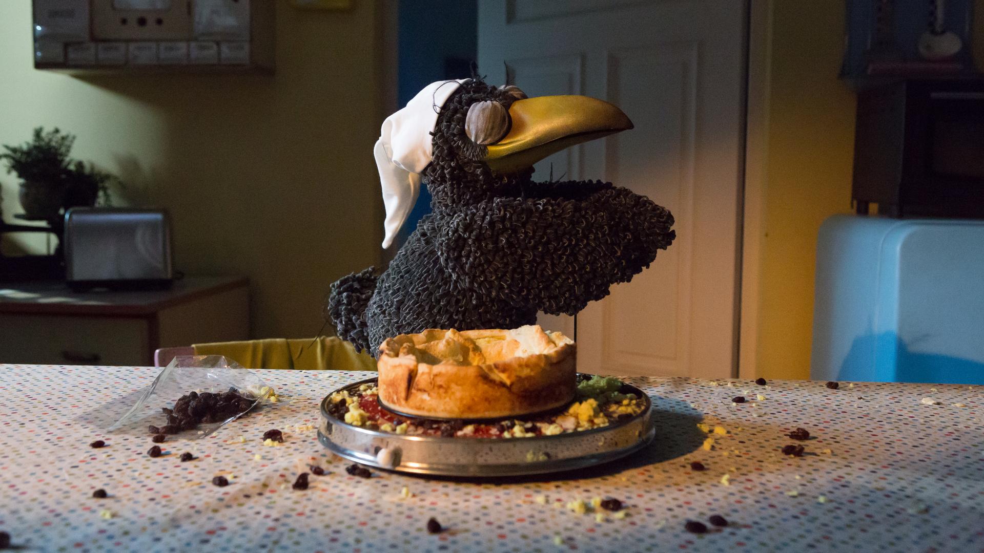 Rudi "schlafwandert" mit geschlossenen Augen und einer Schlafmütze auf dem Kopf neben einem angeknabberten Käsekuchen. Daneben liegen Krümel und Rosinen auf dem Tisch verteilt.