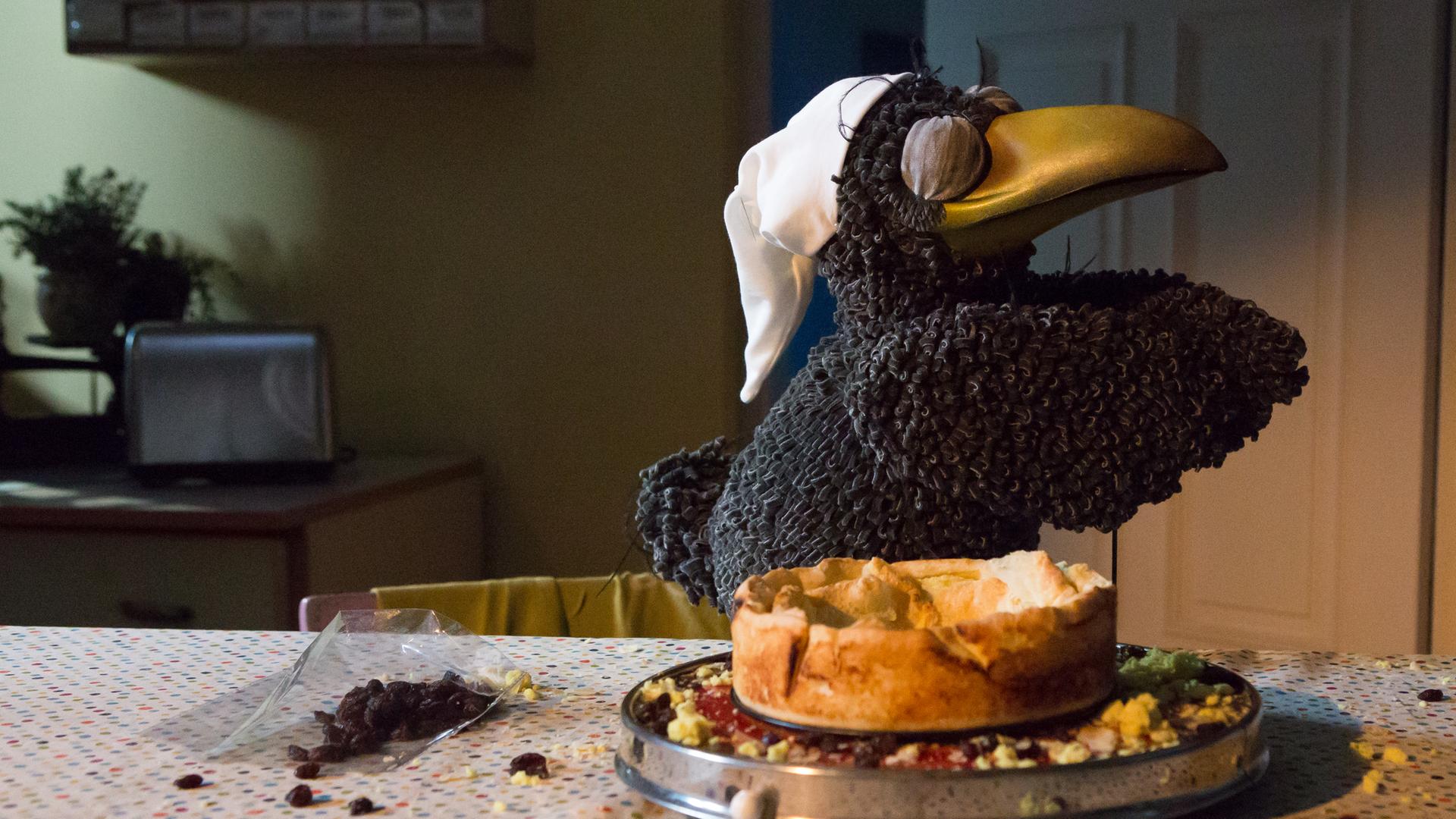 Rudi "schlafwandert" mit geschlossenen Augen und einer Schlafmütze auf dem Kopf neben einem angeknabberten Käsekuchen. Daneben liegen Krümel und Rosinen auf dem Tisch verteilt.