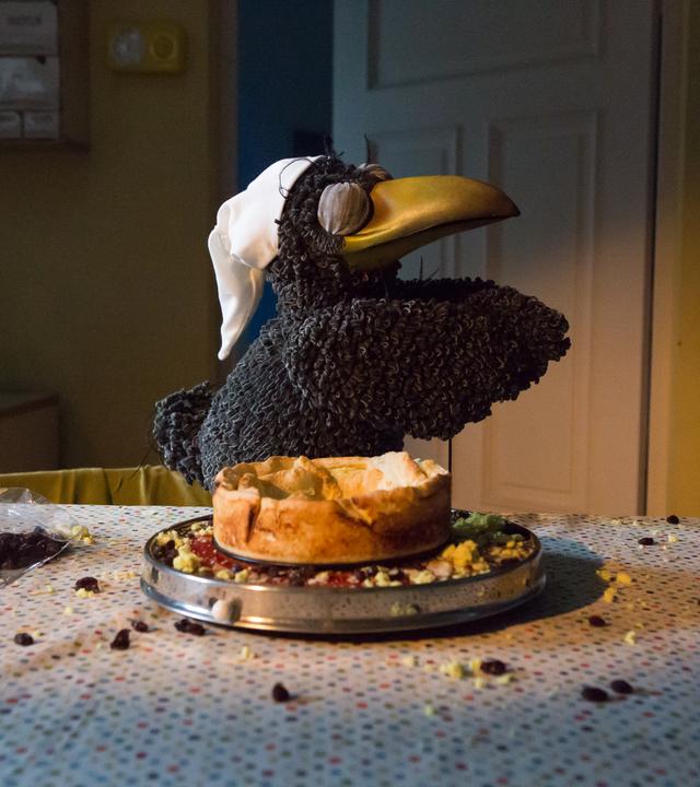Rudi "schlafwandert" mit geschlossenen Augen und einer Schlafmütze auf dem Kopf neben einem angeknabberten Käsekuchen. Daneben liegen Krümel und Rosinen auf dem Tisch verteilt.