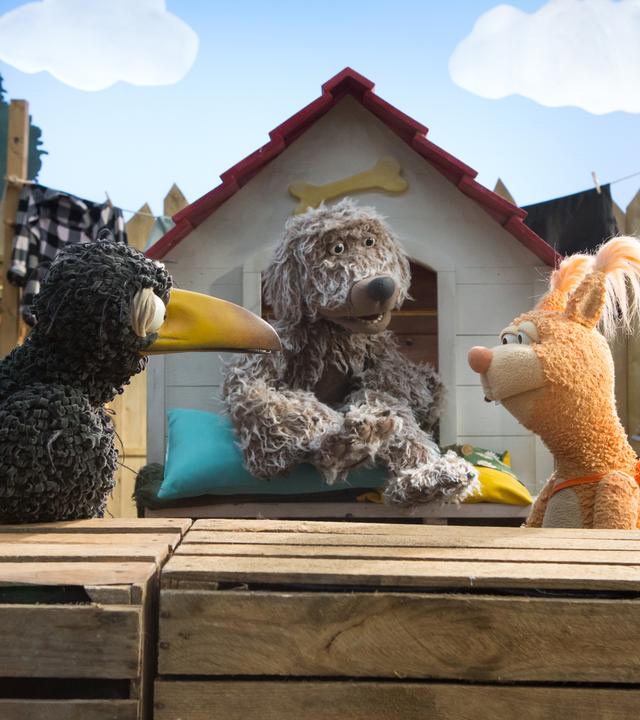 Rabe Rudi und Eichhörnchen Ella stehen sich vor der Hundehütte gegenüber. In der Hütte sitzt Hund Buffo und schaut den beiden zu. Ella hat eine rote Schleife am Schwanz.