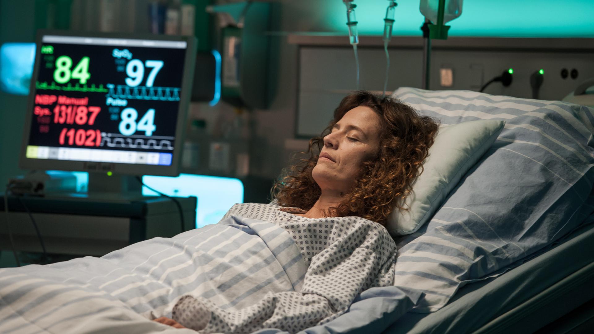 "Der Bergdoktor - Der schlimmste Schmerz (2)": Rike Jäger (Regula Grauwiller) liegt mit geschlossenen Augen im Krankenbett und ist an diverse Apparaturen angeschlossen.