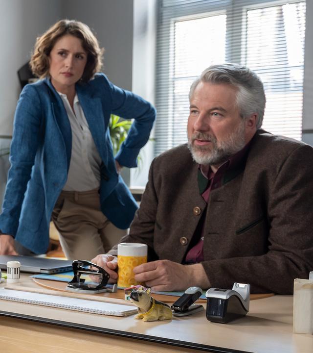 Sarah Weidinger (Kristin Alia Hunold) sitzt auf einem Besucherstuhl vor dem Schreibtisch von Kommissar Stadler (Dieter Fischer), sie hält eine Handtasche auf ihrem Schoß fest. Neben Stadler steht Kommissarin Andresen (Sophie Melbinger), die sich mit der Hand auf die Tischkante stützt und Sarah Weidinger ansieht.