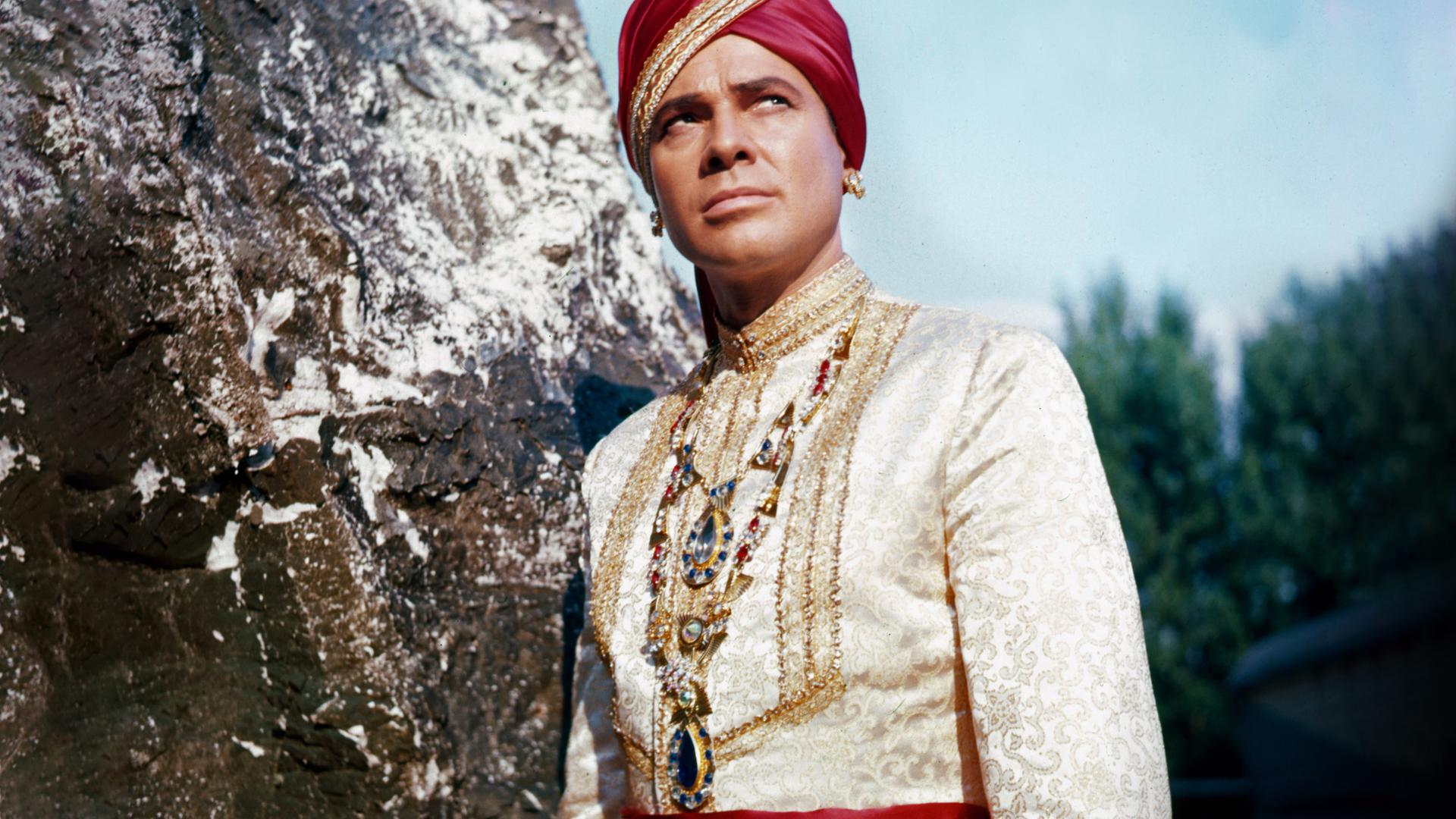 Der Maharadscha von Eschnapur (Walther Reyer) trägt eine herrschaftliche weiße Tracht mit einer prunkvollen Halskette, einer roten Schärpe sowie einem roten Turban und lehnt wie in Pose stehend an einem Felsen. Sein skeptisch-versonnen wirkender Blick richtet sich nach schräg oben.