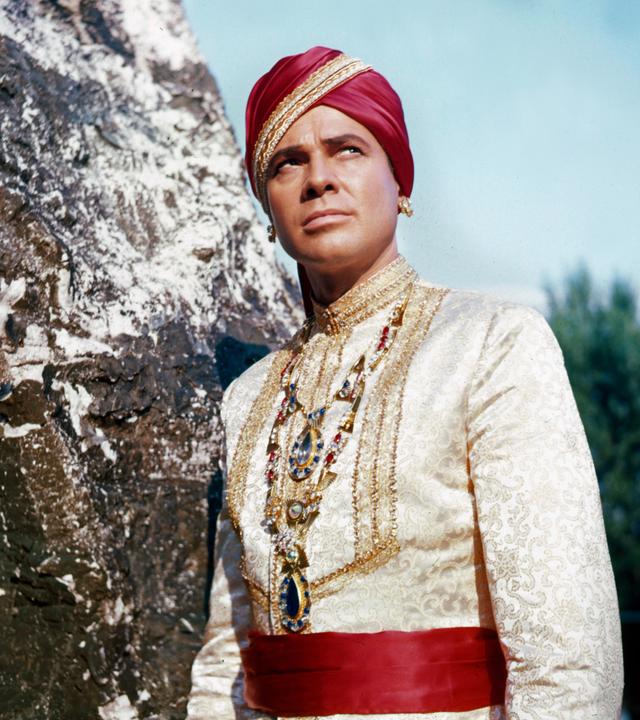 Der Maharadscha von Eschnapur (Walther Reyer) trägt eine herrschaftliche weiße Tracht mit einer prunkvollen Halskette, einer roten Schärpe sowie einem roten Turban und lehnt wie in Pose stehend an einem Felsen. Sein skeptisch-versonnen wirkender Blick richtet sich nach schräg oben.